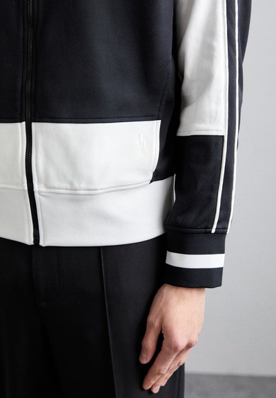 Black and white zippered athletic jacket with contrasting horizontal stripes, smooth fabric, ribbed cuffs, and a subtle logo detail.
