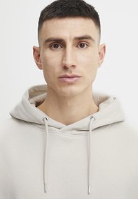 Svetlo béžová mikina vyrobená z mäkkej tkaniny, s šnúrkou na stiahnutie a striebornými agletmi, s voľným strihom a minimalistickým dizajnom.