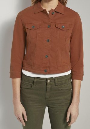 Personne portant une veste boutonnée couleur rouille avec des poches à rabat et un pantalon vert olive, se tenant devant un fond neutre.