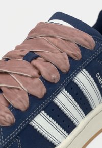 Αθλητικό παπούτσι από σουέτ σε ναυτικό μπλε με χοντρές, ροζ κορδόνια που ξεχωρίζουν. Λευκές δερμάτινες ρίγες στα πλαϊνά και λεπτομέρειες με υφή ραφής.