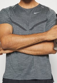 Osoba ubrana w szary, wzorzysty t-shirt sportowy Nike, z założonymi rękami, trzymająca srebrną butelkę wody w prawej ręce.