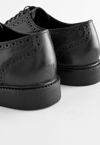 Chaussures en cuir noir avec des détails en brogue, bout arrondi et semelle en caoutchouc noir épaisse avec des accents cousus. Fini texturé sur le dessus.
