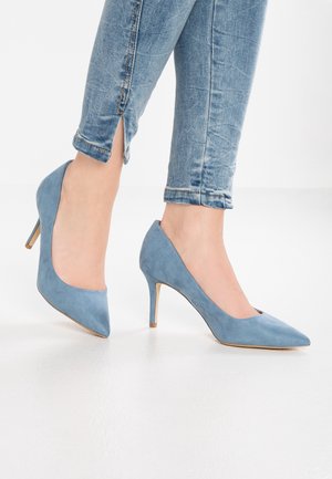 Blå ruskinds højhælede pumps med spidse tæer, der har en slank hæl og en glat tekstur, parret med opfoldede denim jeans.