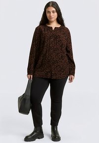 Chemise marron boutonnée avec un motif léopard noir, manches longues, associée à un pantalon noir et des bottines noires, tenant un sac noir.