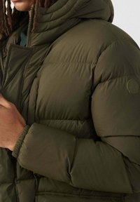 Olijfgroene pufferjack met een hoge kraag, ritssluiting en elastische manchetten. Heeft een gewatteerde textuur en een logo-opdruk op de mouw.