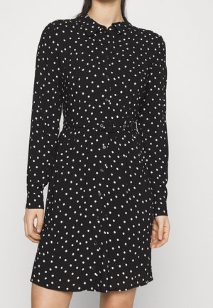 Femme portant une robe noire à manches longues avec des pois blancs, boutonnée devant et une ceinture en tissu assortie à la taille.