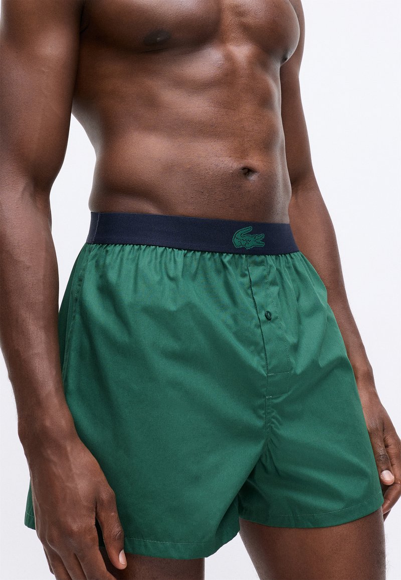 Green swim shorts made of smooth fabric with a navy elastic waistband featuring a dark green logo. Button closure on the front.