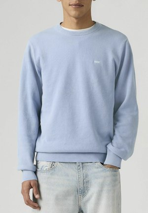 Homme portant un pull à manches longues bleu clair et un jean délavé, avec une main dans la poche et un fond neutre.