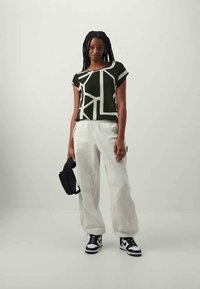 Blusa de mangas curtas com padrão geométrico em preto e branco, combinada com calças largas brancas e sapatilhas pretas, segurando uma pequena bolsa preta.