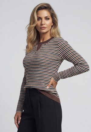 Blondynka z luźnymi falami ma na sobie brązowy, biały i czarny sweter w paski na brązowej koszuli i czarnych spodniach, pozując z jedną ręką na biodrze.