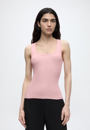 Femme aux longs cheveux foncés portant un haut sans manches rose clair et un pantalon noir, debout devant un fond gris clair uni.