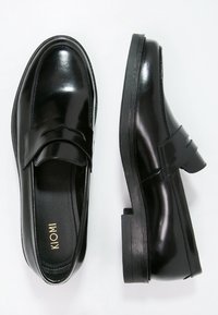Mocassins en cuir noir avec une finition brillante, présentant un design cousu et une semelle plate en caoutchouc texturé. Nom de la marque "KIOMI" visible à l'intérieur.