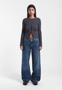 Grijs geribd top met lange mouwen, knoopsluiting en middengedeelte vrijgelaten; gecombineerd met wijde blauwe spijkerbroeken.