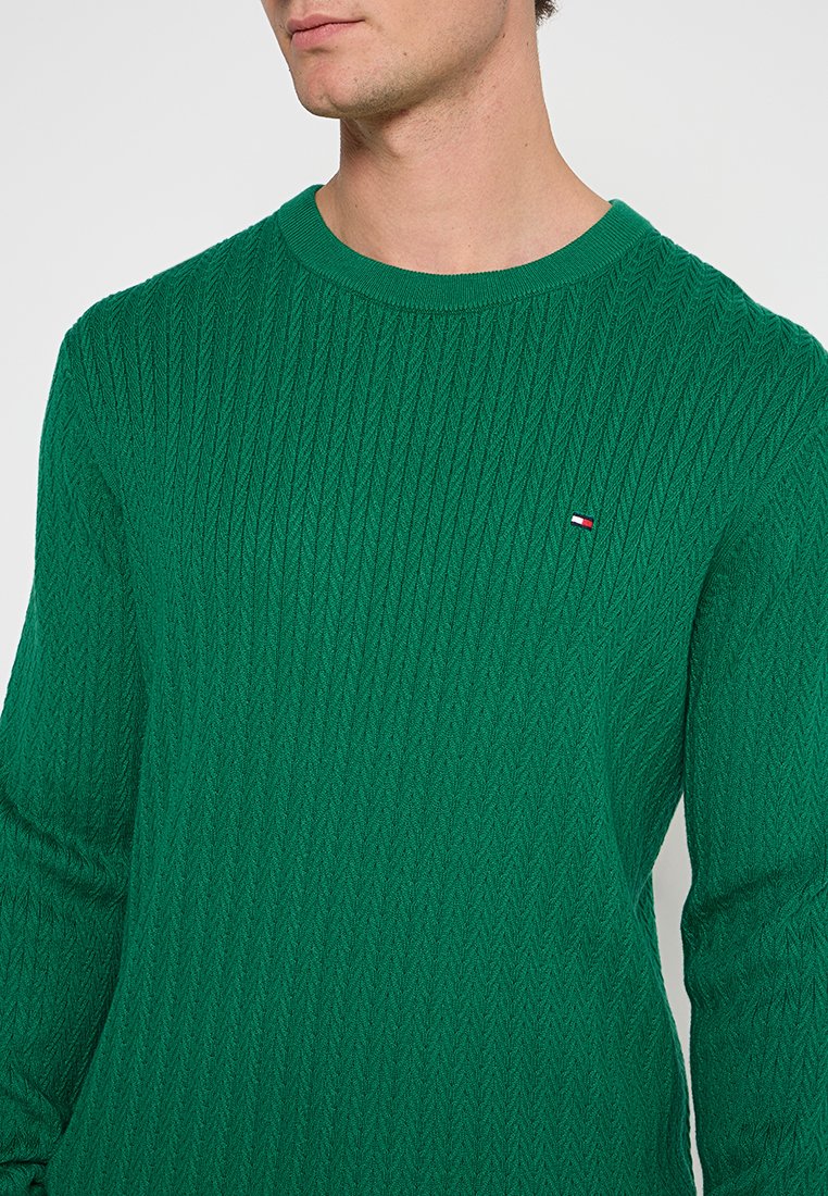 Groene gebreide trui met een gestructureerd ruitpatroon, ronde hals en lange mouwen; heeft een klein logo-accent op de borst.