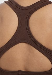 Ruskea urheilullinen hihaton paita, jossa on ristiselkädesign, pehmeä tekstuuri ja ompeluyksityiskohdat, esitelty ihonvärisellä taustalla.