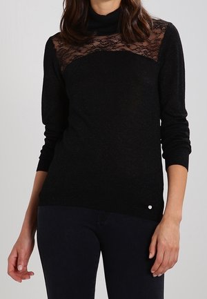 Femme portant un haut noir à manches longues avec un empiècement en dentelle et un col roulé, assorti à un pantalon noir ajusté, sur un fond uni.