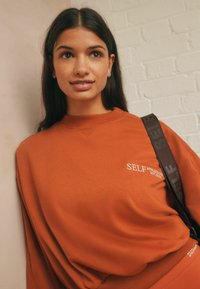 Huppari terrakottavärissä, joissa on ribbireunuskaulus, ja jossa on valkoiset painatukset "SELF ATHLEISURE EST. 2023." Pehmeää kangasta, rento istuvuus.