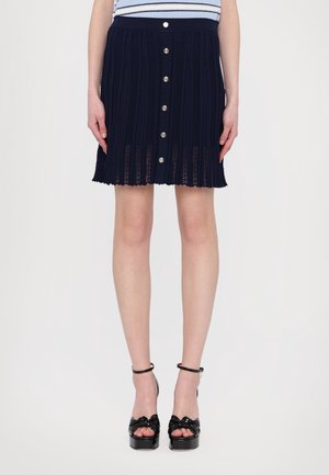 Vrouw draagt een marineblauwe gebreide rok met zilveren knopen aan de voorkant, gecombineerd met zwarte sandalen met hoge hakken en enkelbandjes.