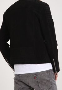 Giacca in suede nera con pannelli a coste sulle spalle e cerniere laterali. Indossata sopra una camicia bianca con jeans grigi.