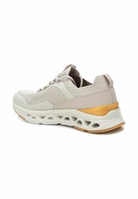 Zapatilla deportiva color beige claro con parte superior de malla, acentos texturizados y una suela blanca acolchada que cuenta con un acento dorado en el talón.