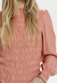 Blushfärgad smockad topp med långa ärmar, med ett strukturerat tyg och samlade detaljer som skapar ett varierat ytmönster.