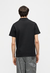 Άνδρας που στέκεται με την πλάτη στην κάμερα, φοράει απλό μαύρο πόλο με κοντό μανίκι και μεταλλικά γκρι παντελόνια με μικρό μαύρο λογότυπο.