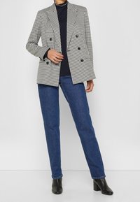 Vrouw in een zwart-wit houndstooth dubbelgebreide blazer, zwarte top, blauwe jeans en zwarte haklaarzen tegen een witte achtergrond.
