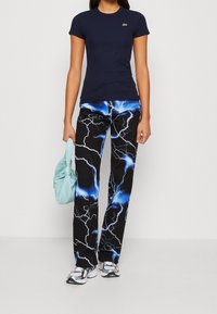 Žena oblečená v námořnickém tričku s krátkým rukávem, černých kalhotách s modrým bleskovým potiskem, bílých teniskách a držící světle modrou kabelku.