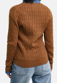 Brauner gestrickter Pullover mit einem strukturierten Zopfdesign, der über einem blauen Kragenshirt getragen wird. Der Pullover hat einen gerippten Saum und Bündchen.