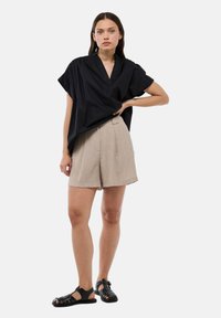 Haut noir ample à manches courtes avec un col en V, associé à un short beigne plissé et des sandales noires à lanières, sur fond blanc.