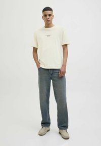 Cremefarvet bomulds T-shirt med korte ærmer og trykt tekst på forsiden. Bæres med lyseblå jeans og beige sko.