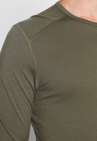 Cămașă cu mânecă lungă de culoare verde măslinie, fabricată dintr-un material moale și texturat, cu cusături contrastante pe laturile umerilor.