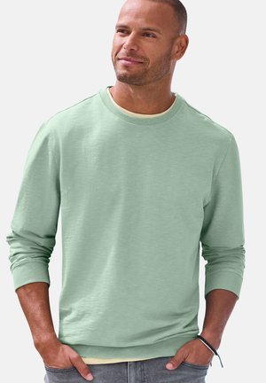 Mann mit kurzen Haaren und Bart, trägt ein hellgrünes Sweatshirt mit langen Ärmeln, ein hellgelbes Hemd darunter und graue Hose, die Hände in den Taschen.