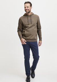 Hnedá bavlnená mikina s prednou kapsou a modrým potlačeným textom. K outfitu dopĺňajú úzke námořnícke nohavice a čierne topánky.