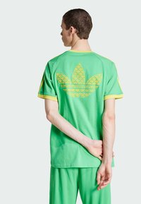 Zöld pamut póló, nagy sárga mintás Adidas logóval a hátán, amelyet sárga díszítések egészítenek ki az ujjaknál.