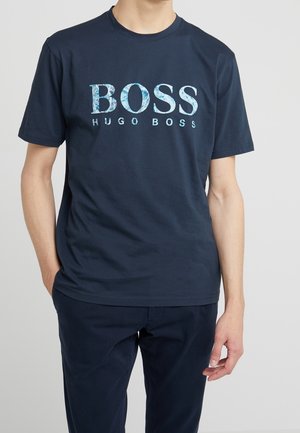 Osoba ubrana w granatowy t-shirt Hugo Boss z napisem „BOSS HUGO BOSS” w jaśniejszym niebieskim wzorze, zestawiony z ciemnoniebieskimi spodniami.