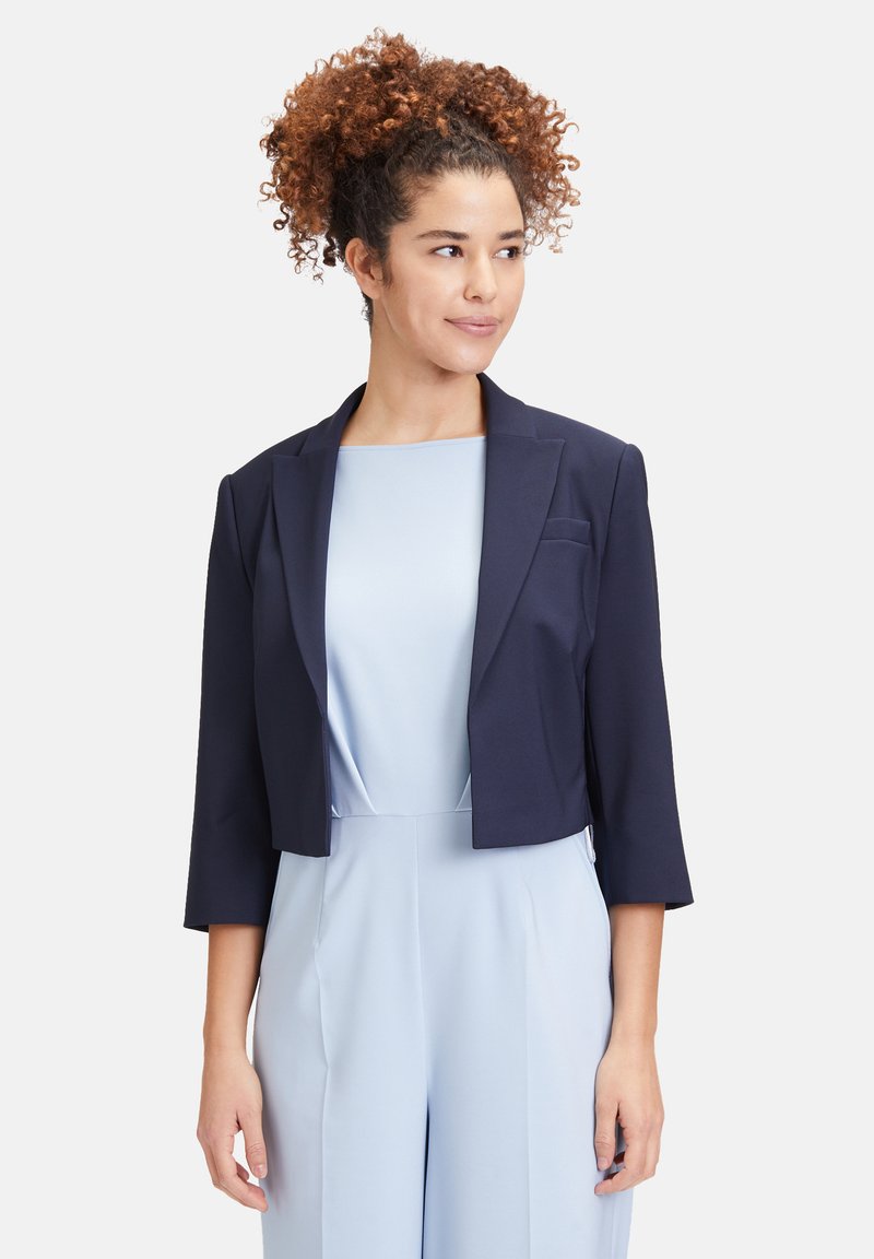 Donkerblauwe cropped blazer met driekwartmouwen, gladde stof, slanke vormgeving, reverskraag en een enkele borstzak. Gecombineerd met een lichtblauwe jumpsuit.