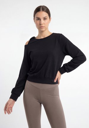 Junge Frau mit dunklen Haaren trägt ein schwarzes Langarmshirt mit Schulterauschnitt und taupefarbene Leggings, posiert vor weißem Hintergrund.