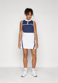 Camisa polo de color navy con cuello y rayas blancas, combinada con una falda blanca con acentos navy en los laterales. La tela es suave y tiene un diseño deportivo.