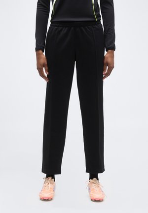 Persoană care poartă pantaloni sport negri cu talie elastică și adidași sportivi piersică, stând pe un fundal deschis simplu.