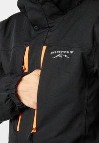 Zwarte geïsoleerde jas met oranje ritsaccenten en een logo. Kenmerkt zich door een hoge kraag, verstelbare manchetten en ritszakken aan de voorkant.