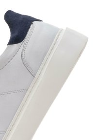 Sneaker blanche avec un dessus en cuir lisse, un talon en daim bleu marine et une semelle en caoutchouc blanche. Présente des détails de couture sur le côté.