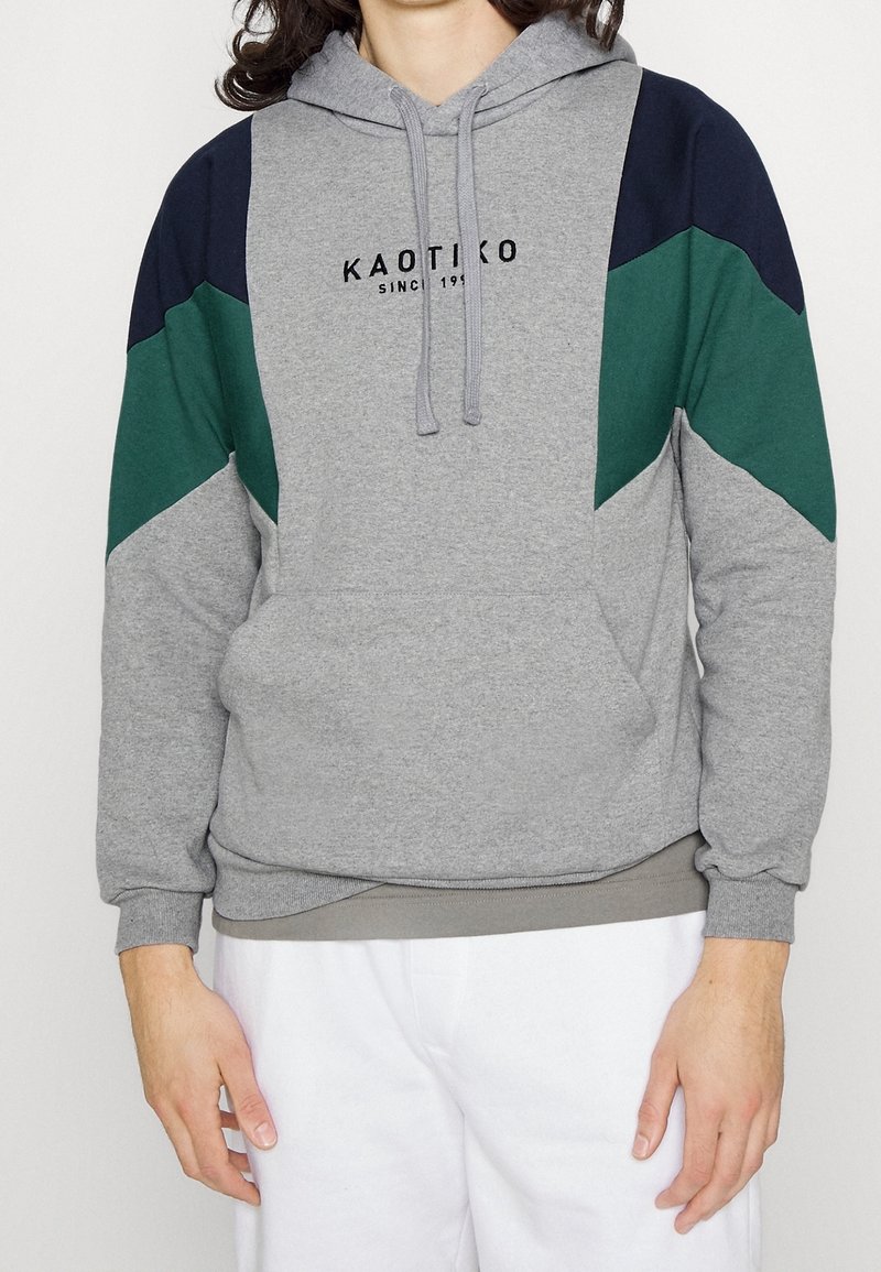 Personne portant un sweat à capuche gris avec des patches d'épaules bleu marine et verts et le texte "KAOTIKO Since 1993", associé à un pantalon blanc.