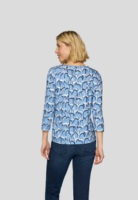 Blau gemustertes Oberteil mit Dreiviertelärmeln, das ein wellenförmiges Design in unterschiedlichen Blau- und Weißtönen aufweist. Getragen mit dunkler Denim.