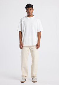 Un joven se encuentra de pie mirando hacia adelante, vistiendo una camiseta blanca oversized sencilla, pantalones de color blanco sucio y zapatillas de color claro, contra un fondo blanco.