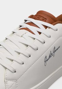 Λευκό αθλητικό παπούτσι με λευκά κορδόνια, καφέ γλώσσα και το λογότυπο "Jack & Jones" σε μαύρο χρώμα στο πλάι.