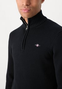 Mann iført en svart ribbestrikket genser med høy hals, med en sølvglidelås som er delvis åpen, og en brodert GANT-logo på brystet.