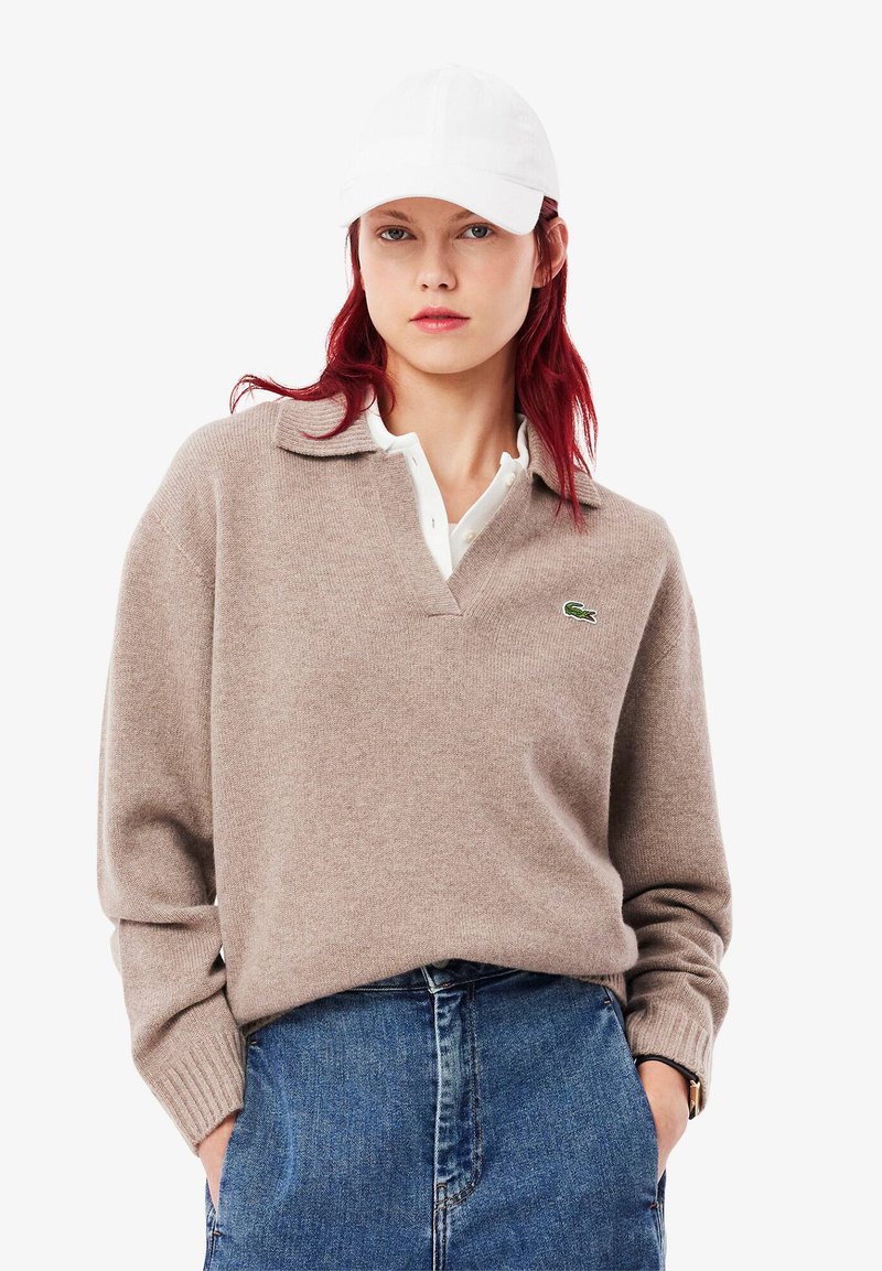 Beiger Strickpullover mit V-Ausschnitt und gerippten Bündchen, über einem weißen Hemd mit Kragen getragen. Verfügt über ein kleines grünes Logo auf der linken Brustseite.