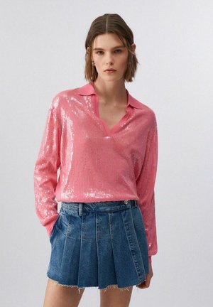 Donna che indossa una maglia rosa lucida a maniche lunghe con colletto e una minigonna di jeans blu plissettata, mani nelle tasche, sfondo neutro.