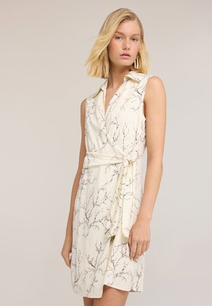 Blonde Frau mit kurzen Haaren trägt ein ärmelloses cremefarbenes Wickelkleid mit feinem Zweigmuster und großen Creolen, steht vor einem schlichten Hintergrund.